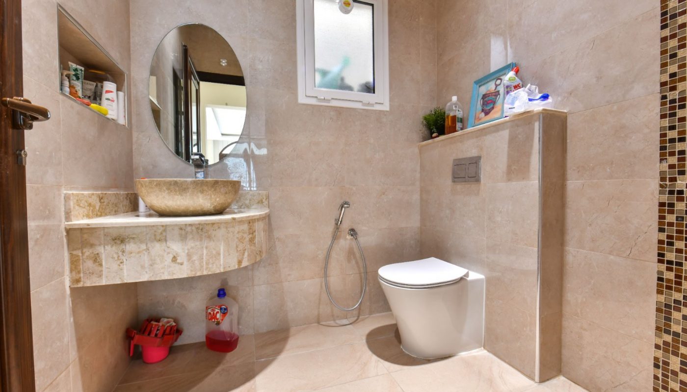 A modern bathroom with beige tiles, featuring a circular mirror, a bowl sink, and a wall-mounted toilet. A small shelf holds toiletries, and a bidet spray is next to the toilet.