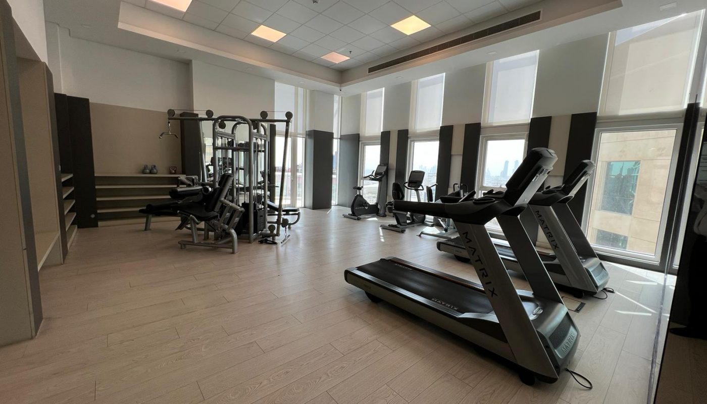 A modern gym with treadmills, stationary bikes, a multi-station weight machine, and shelves with towels, all in a well-lit room with large windows.
