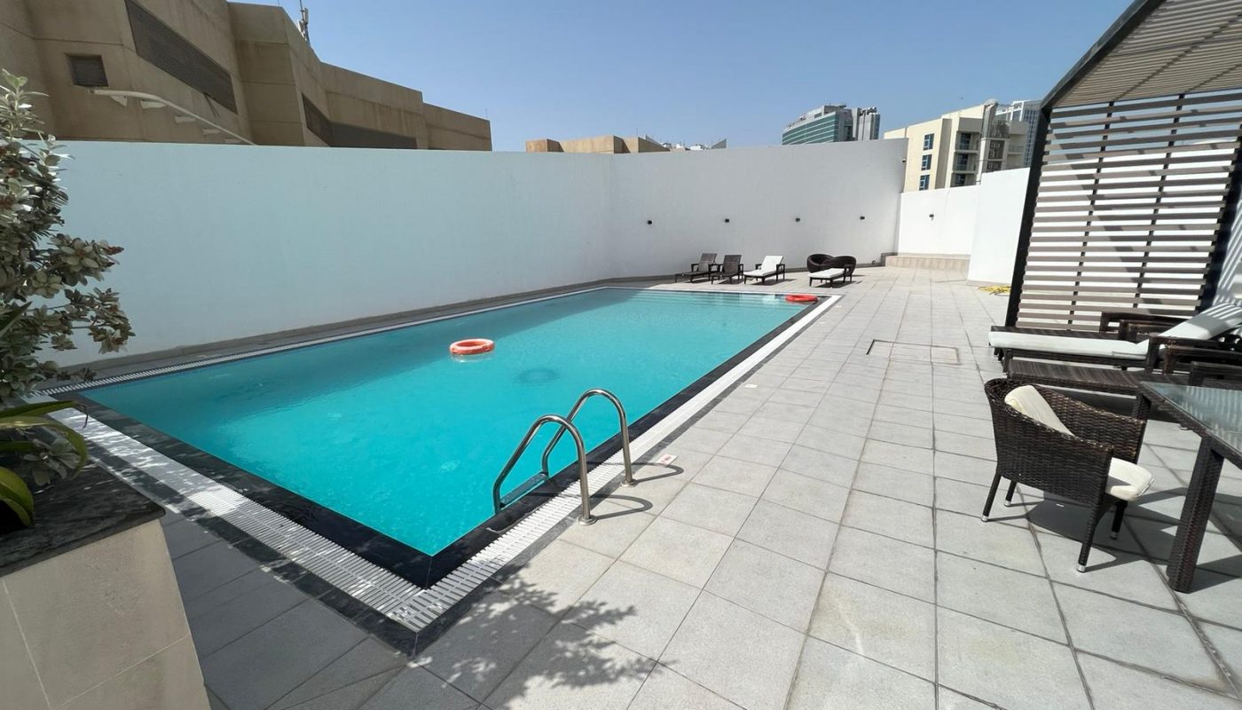 Rectangular outdoor swimming pool with a ladder, lifebuoy, lounge chairs, and shaded seating area on a tiled patio, surrounded by tall white walls and buildings.