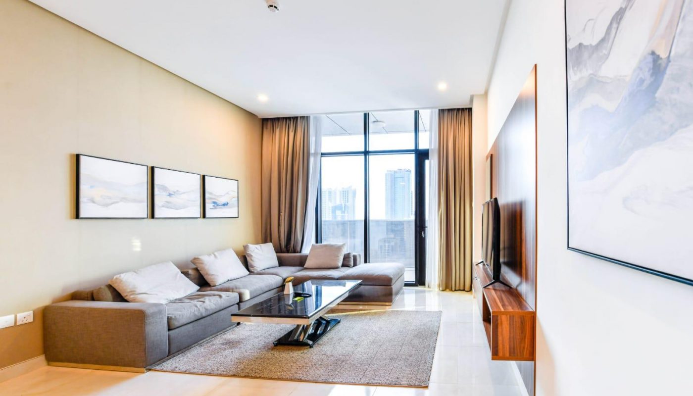 Modern living room with a gray sectional sofa, wall art, a rug, a coffee table, TV on a wooden panel, large windows, and city skyline view.