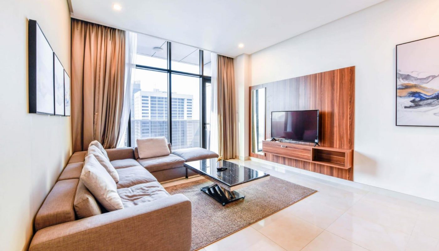 Modern living room with a sectional sofa, glass coffee table, wall-mounted TV on wood panel, large window with city view, and neutral-colored decor.