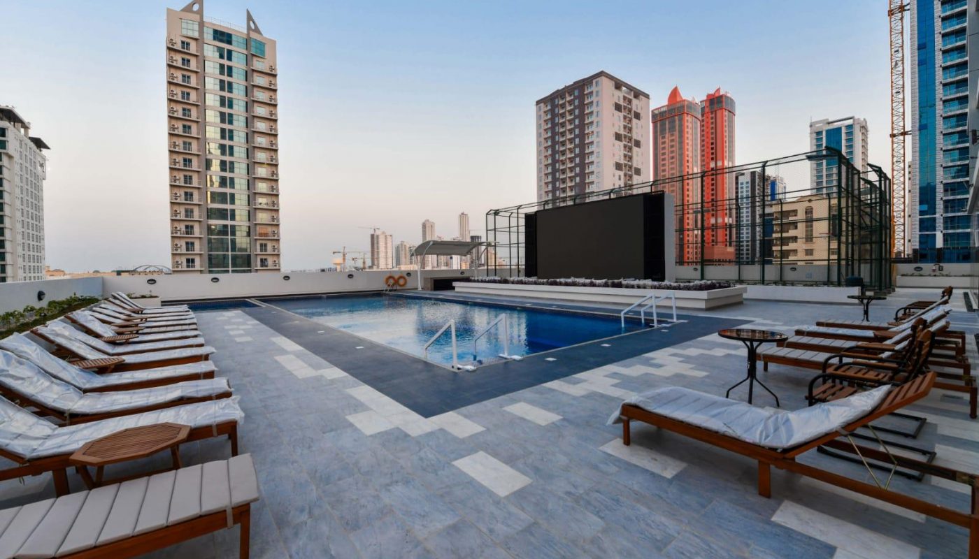 Rooftop pool area with lounge chairs, a large screen, and high-rise buildings in the background during daytime.
