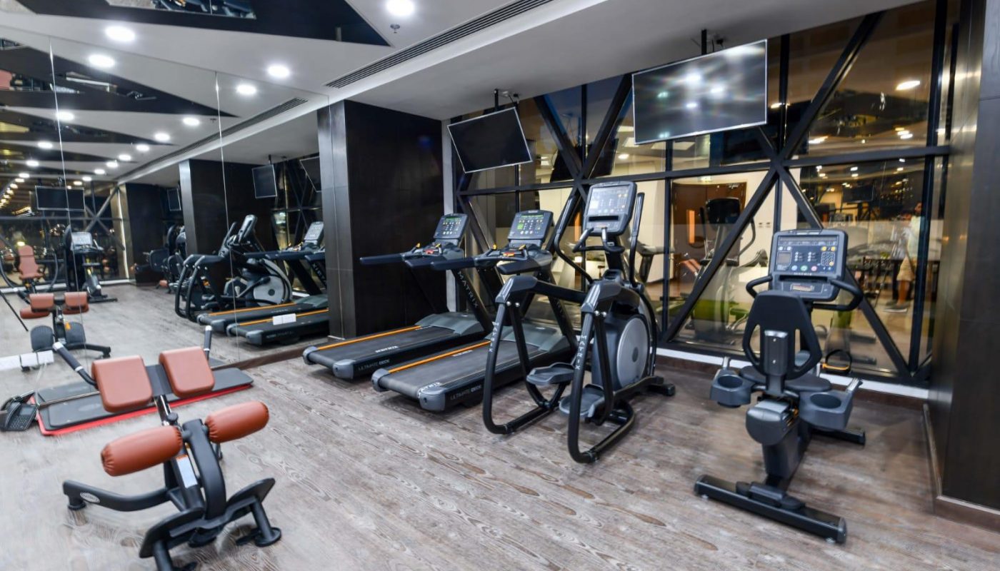 Modern gym with treadmills, exercise bikes, and weight benches on wooden flooring, large mirrors on the wall, and TVs mounted above the cardio machines.