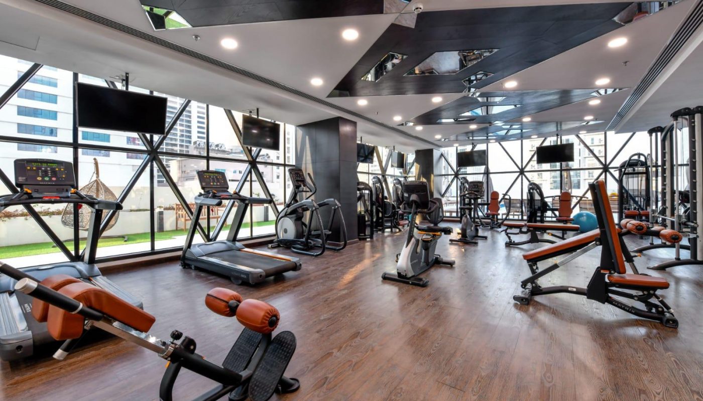Modern gym with wooden floors, large windows, cardio machines, weight benches, and strength equipment in a well-lit space.