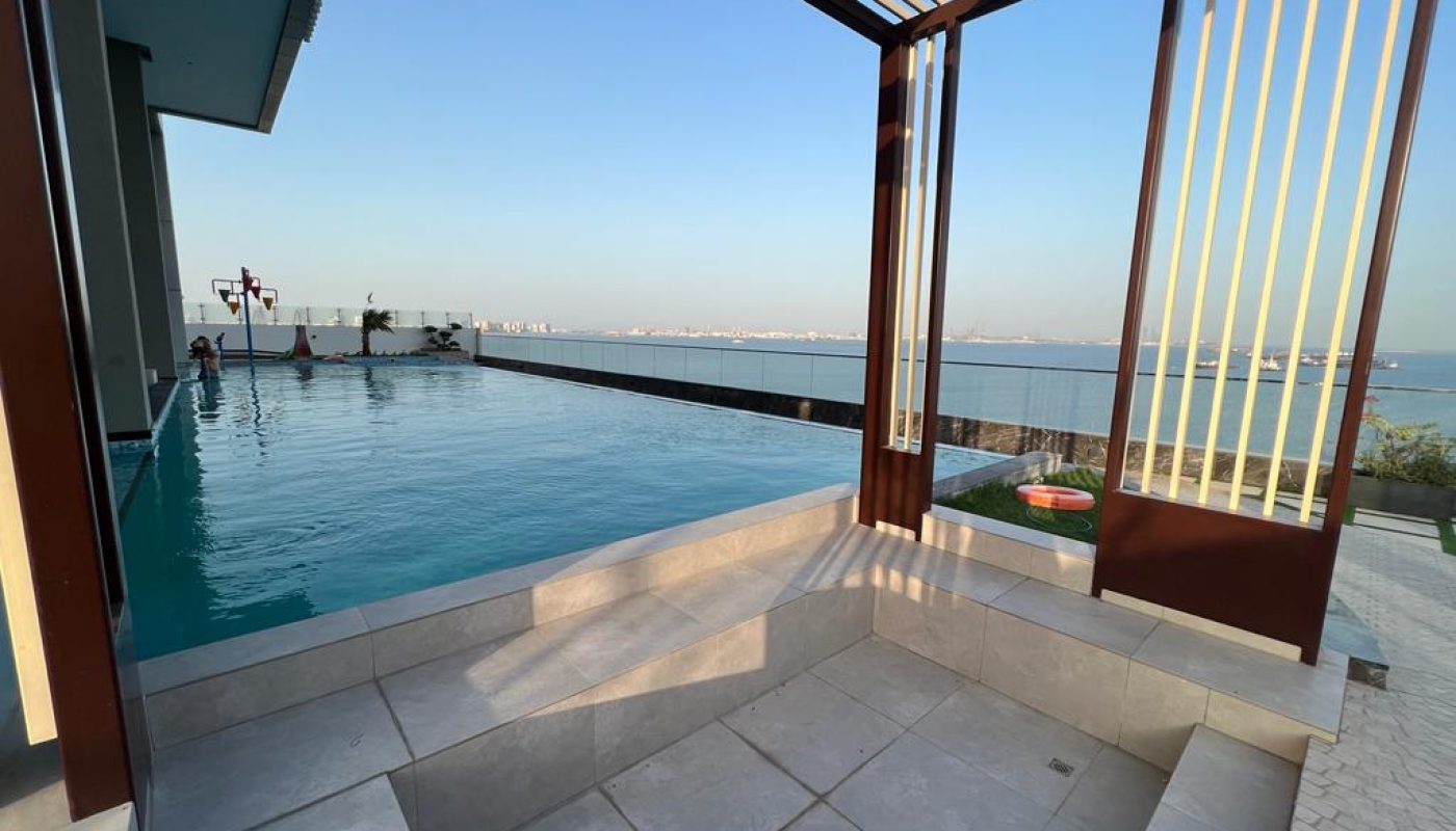 Infinity pool with a shaded seating area overlooking the sea, under a clear sky, with a distant cityscape and ships visible on the horizon.