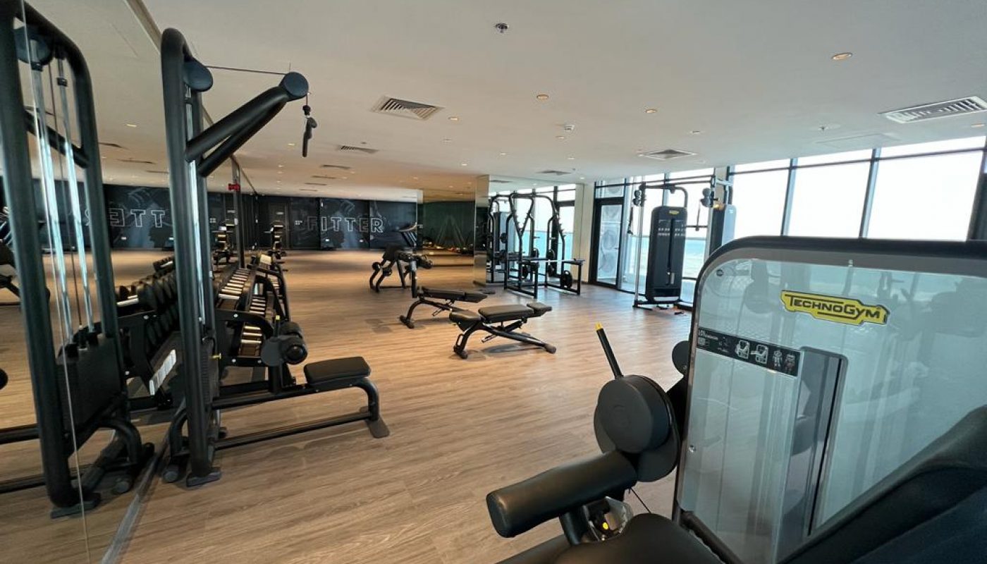 Spacious gym with various exercise machines, benches, and mirrored walls. Large windows provide natural light, and the room has wood-style flooring.