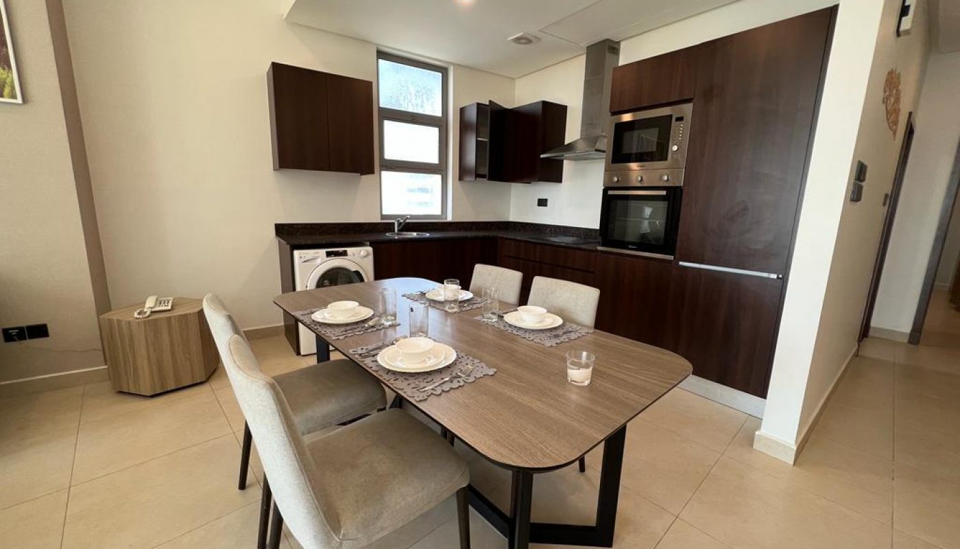 Modern kitchen with dark wood cabinets, built-in appliances, a washing machine, and a dining table set for four with neutral-colored chairs.
