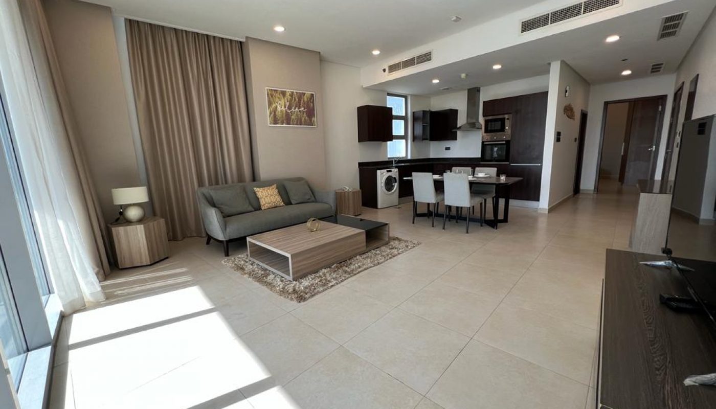 Modern open-plan living room and kitchen with neutral colors, tiled floor, sofa, dining table, wall art, and large windows letting in sunlight.