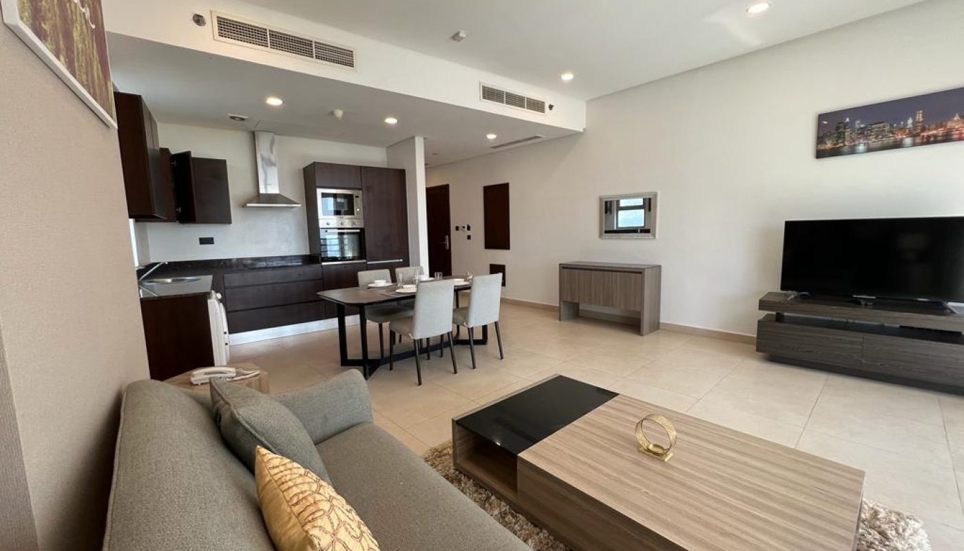 Modern open-plan living area with a gray sofa, coffee table, dining set, flat-screen TV, and kitchen with dark cabinets. Neutral colors and minimal decor.