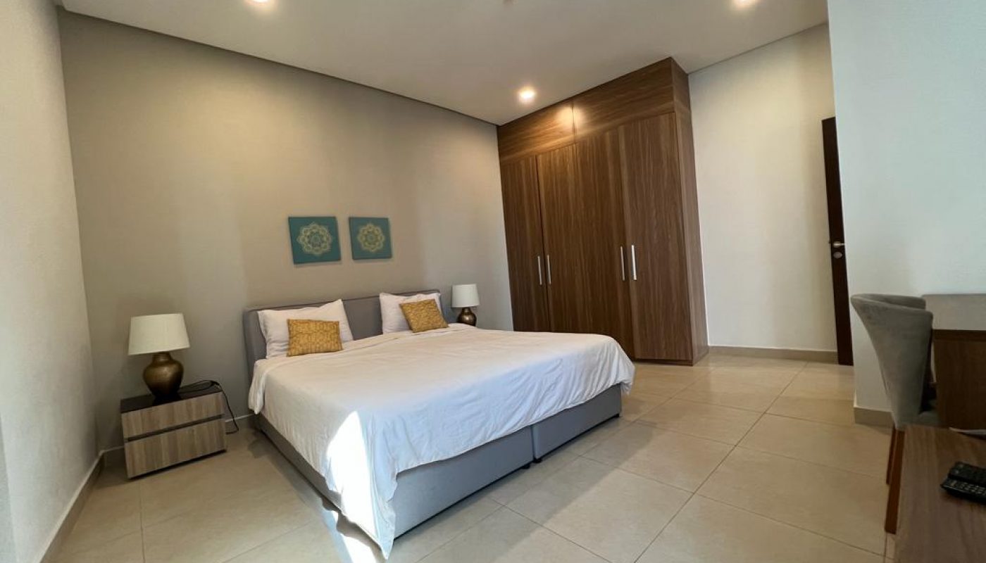 A modern bedroom with a double bed, two bedside tables with lamps, a large wooden wardrobe, and a desk with a chair. The room has tiled floors and neutral-colored walls.