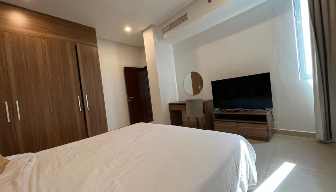 A modern bedroom with a double bed, wooden wardrobe, wall-mounted TV, dressing table with mirror, and a window letting in natural light.