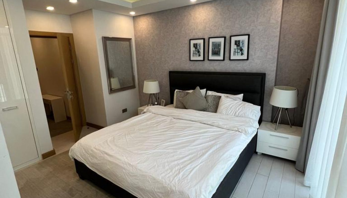 Modern bedroom with a neatly made double bed, two bedside tables with lamps, three framed pictures above the headboard, and a large window with sheer curtains.