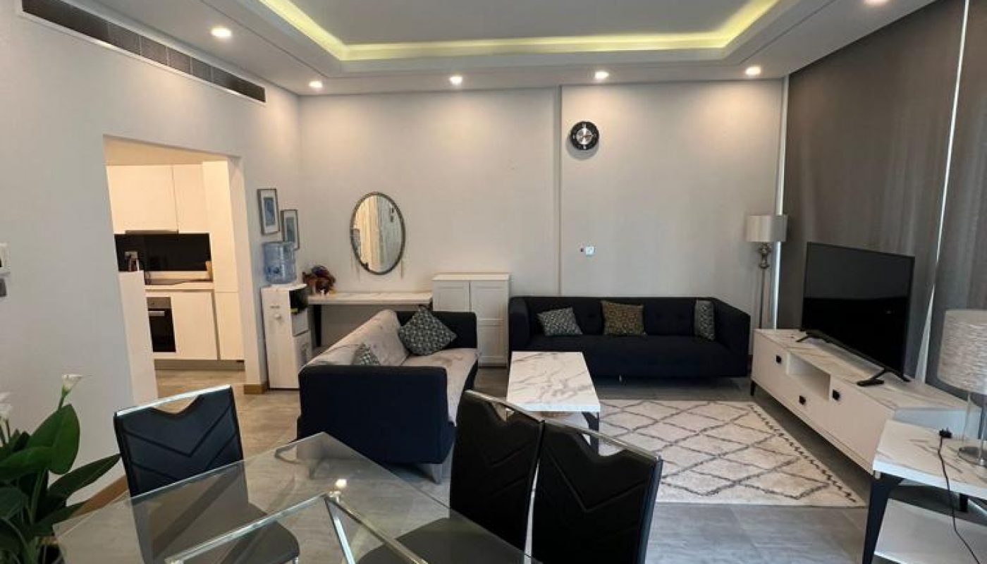 Modern living and dining area with glass dining table, black chairs, two sofas, TV, and neutral decor. Kitchen visible in the background. Ceiling lights and large windows provide illumination.