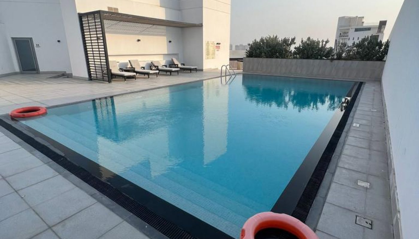 Rooftop swimming pool with clear water, surrounded by white tiles, two orange life buoys, and lounge chairs in the background on a sunny day.