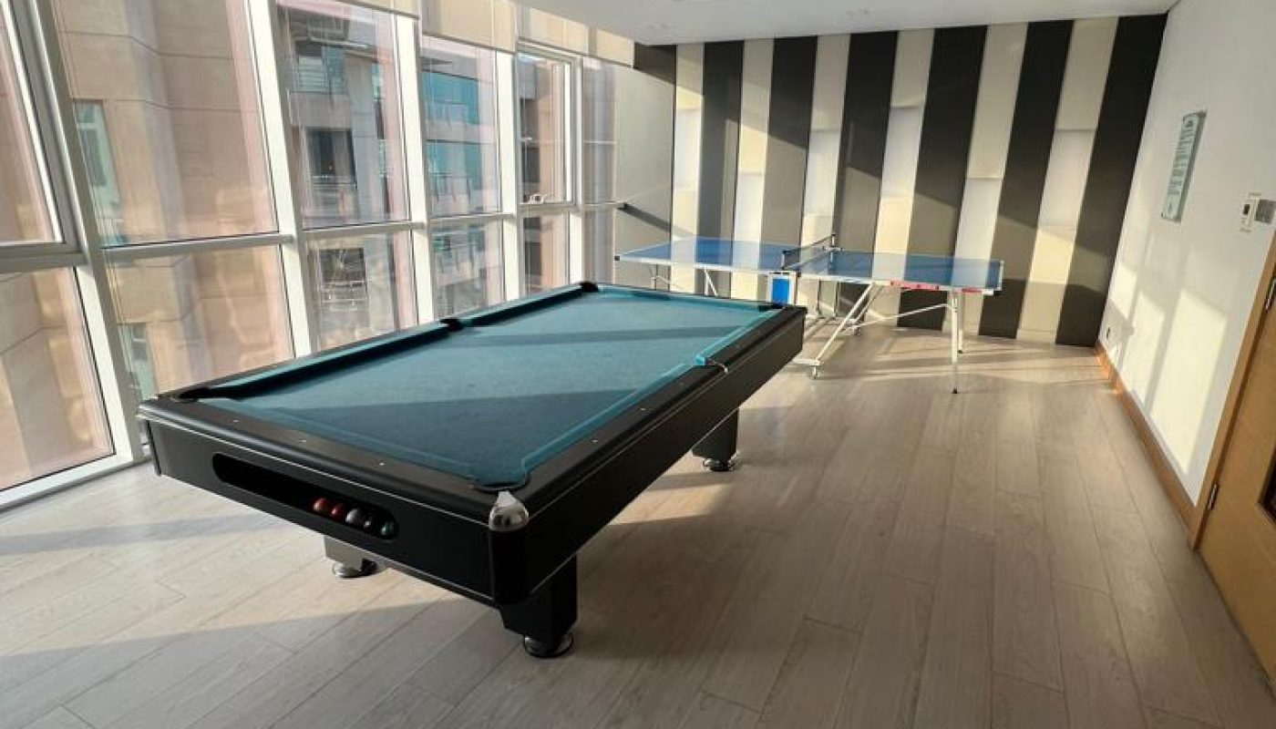 A room with a pool table in the center and a ping pong table by the striped wall, featuring large windows and light wood flooring.