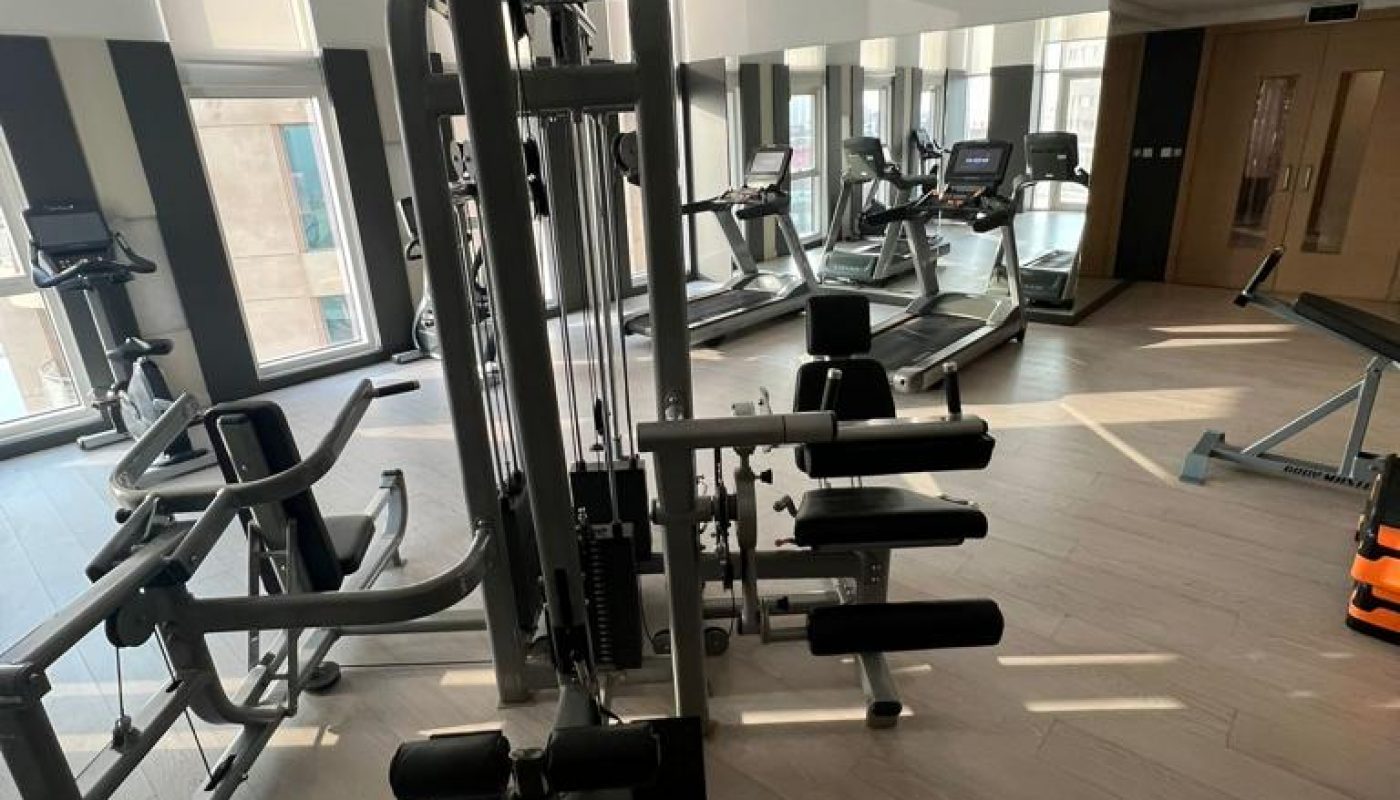 A gym room with strength training equipment, treadmills, ellipticals, and large windows letting in natural light.