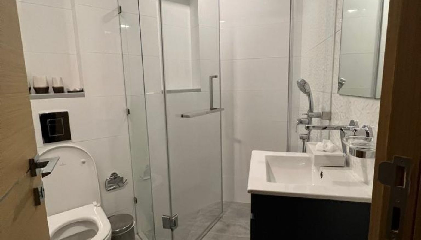 Modern bathroom with a wall-mounted toilet, glass-enclosed shower, and a sink with a black vanity. The floor is gray tile, and the walls are white.