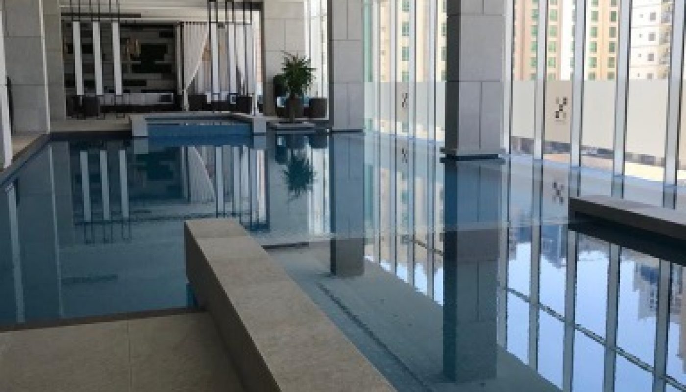 Indoor swimming pool with large windows, city view, and modern decor; a hot tub area is in the foreground, and poolside lounge chairs are visible in the background.