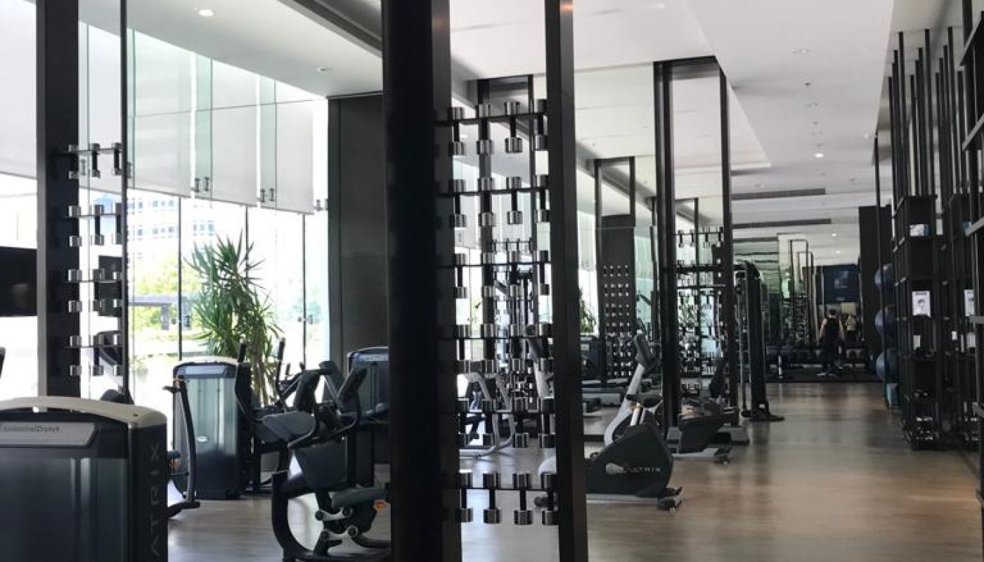 Modern gym with large mirrors, cardio machines, exercise bikes, and natural light coming through tall windows.