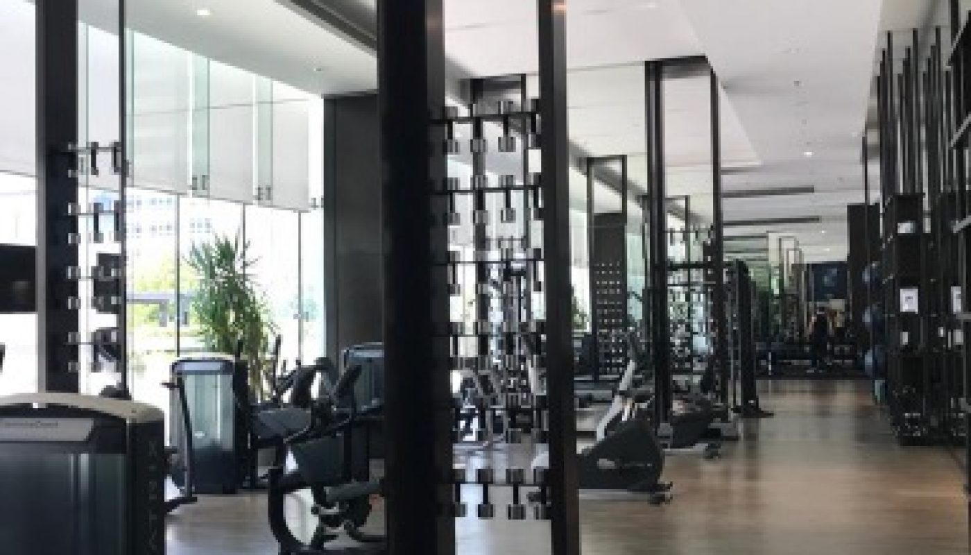 Modern gym with cardio and strength equipment, large mirrors, glass walls, wooden flooring, and natural light from windows.