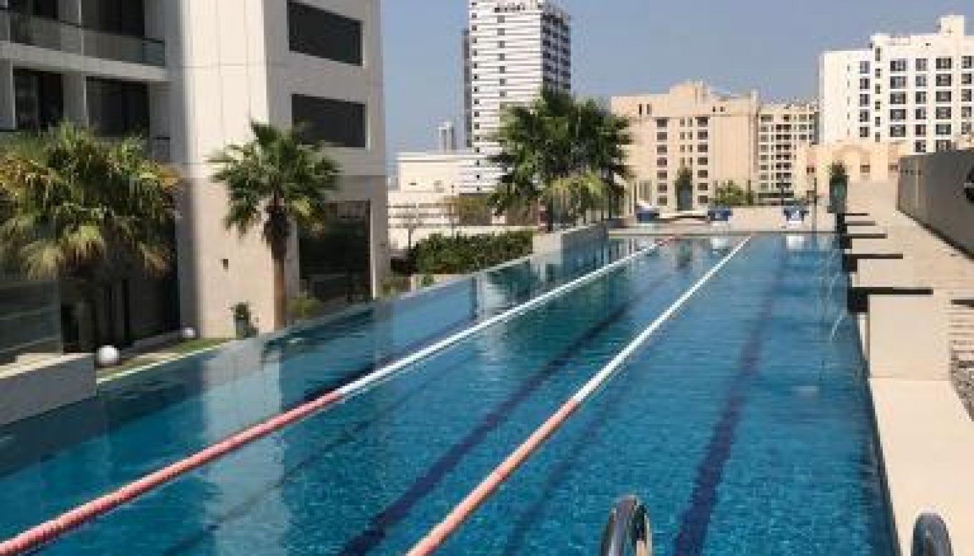 A rooftop swimming pool with lane markers, surrounded by palm trees and tall buildings under a clear blue sky.