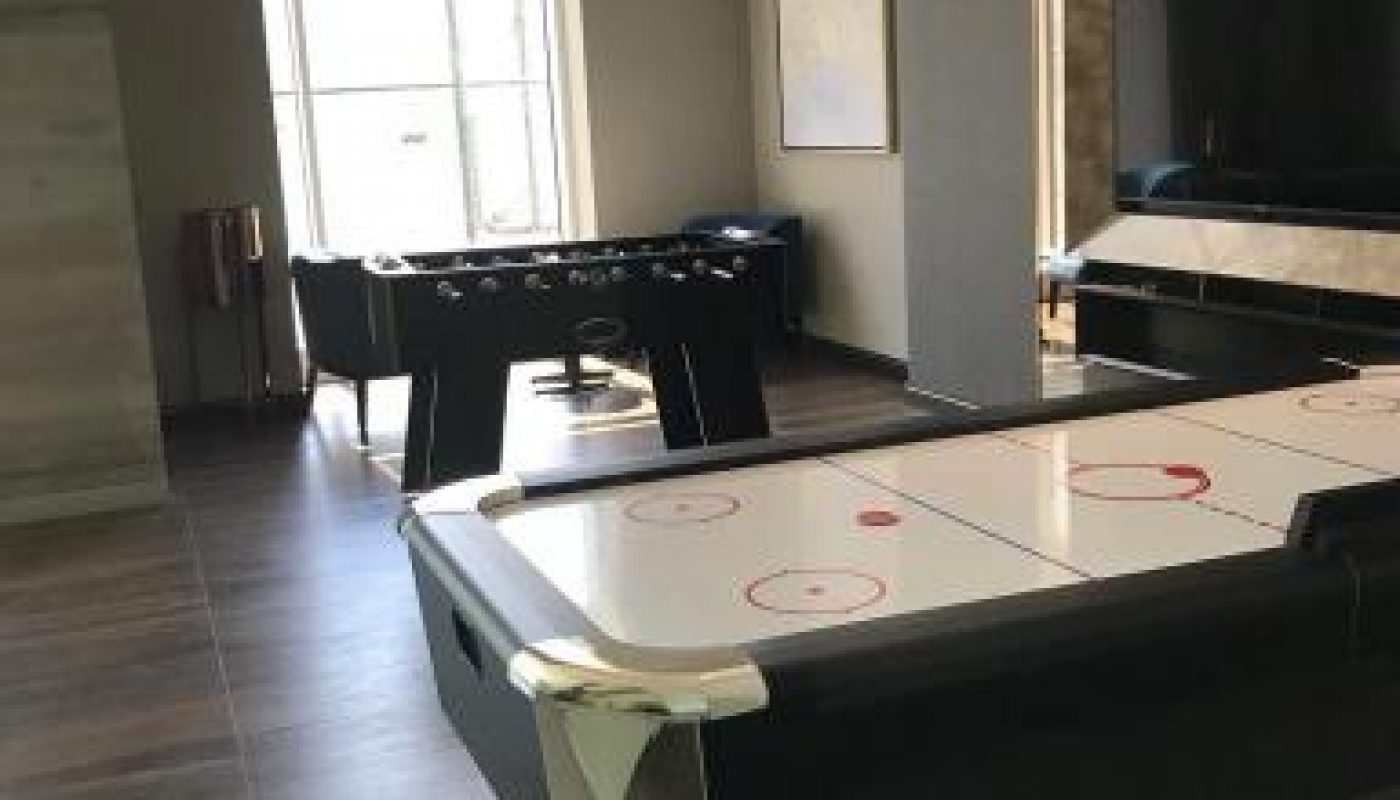 A game room with an air hockey table in the foreground and a foosball table near large windows in the background.