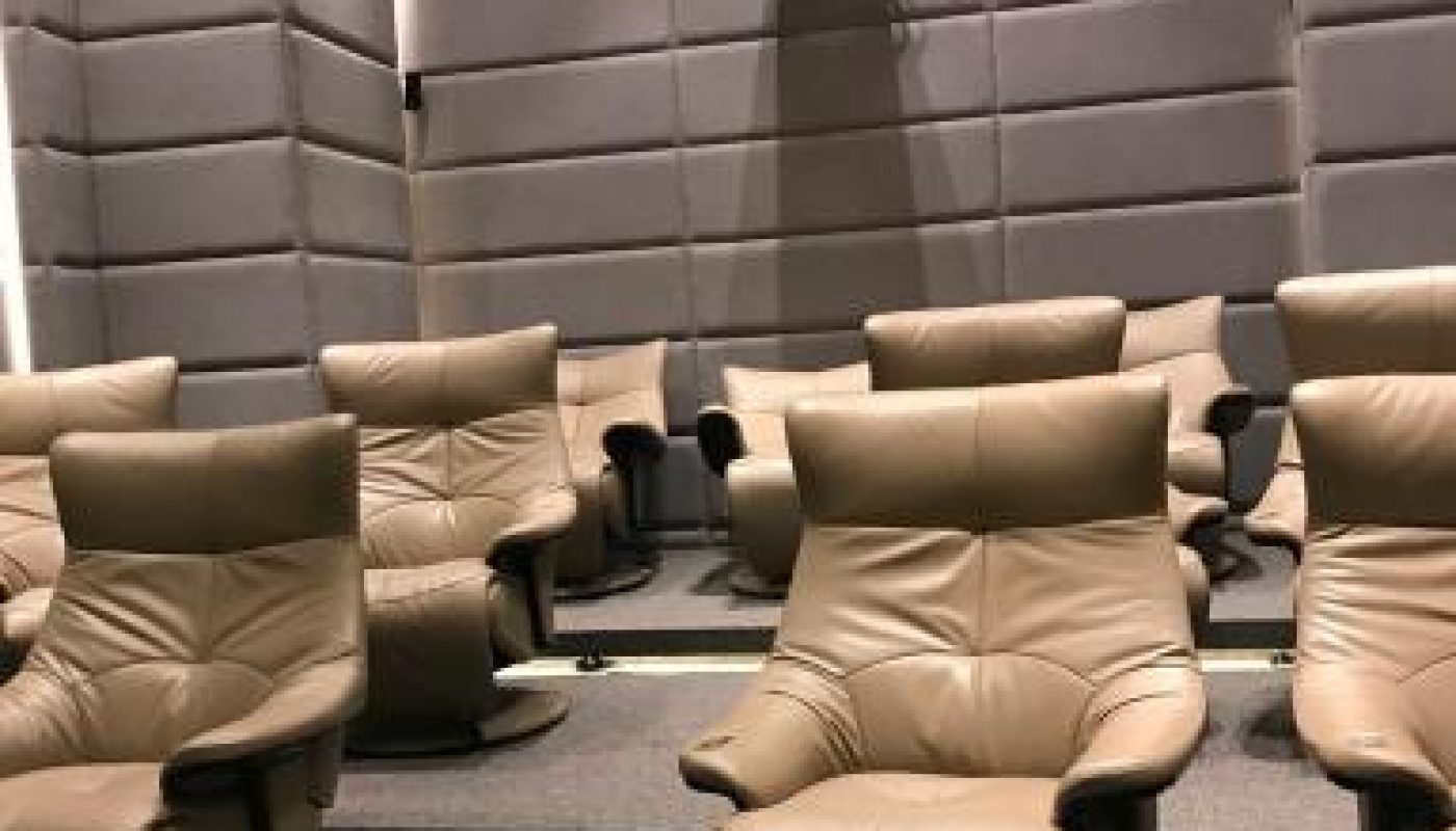 A small theater room with beige leather recliner chairs arranged in rows on tiered carpeted flooring and a projector mounted on the back wall.