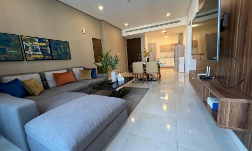 Modern living room in a luxury 2BR apartment with a grey sectional sofa, colorful cushions, and glossy white floor tiles, extending to an open-plan kitchen.