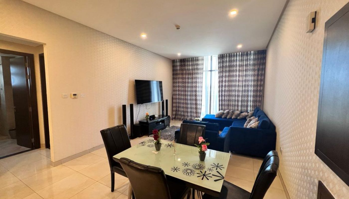 A modern living and dining area with a glass dining table and four chairs, a blue sofa set, a TV on a stand, and floor-to-ceiling patterned curtains.