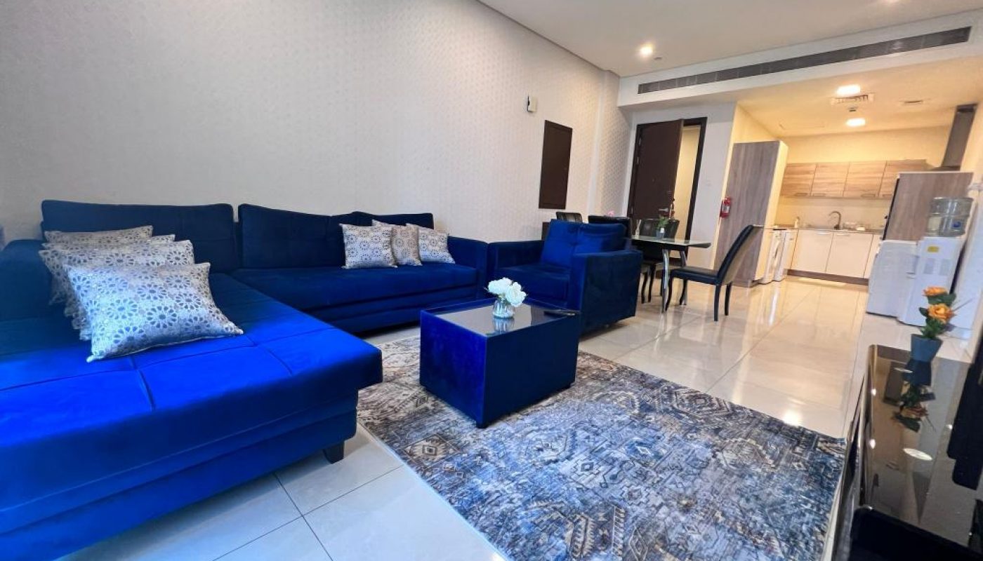Modern living room with blue sofas, a patterned rug, and a coffee table; dining area and kitchen are visible in the background.