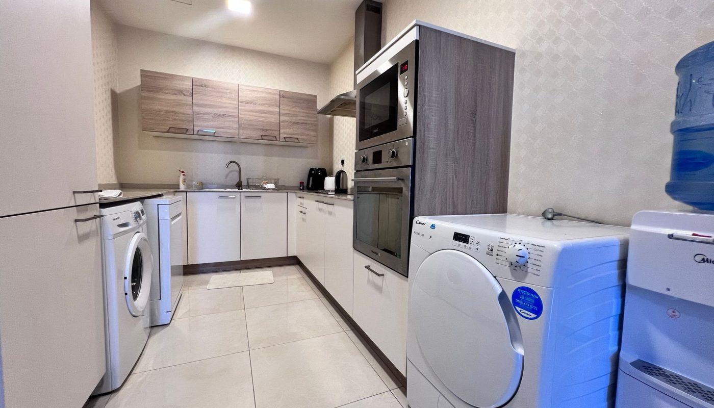 Modern kitchen with white cabinets, built-in oven, washing machine, dryer, sink, countertop appliances, and a water dispenser on the right.