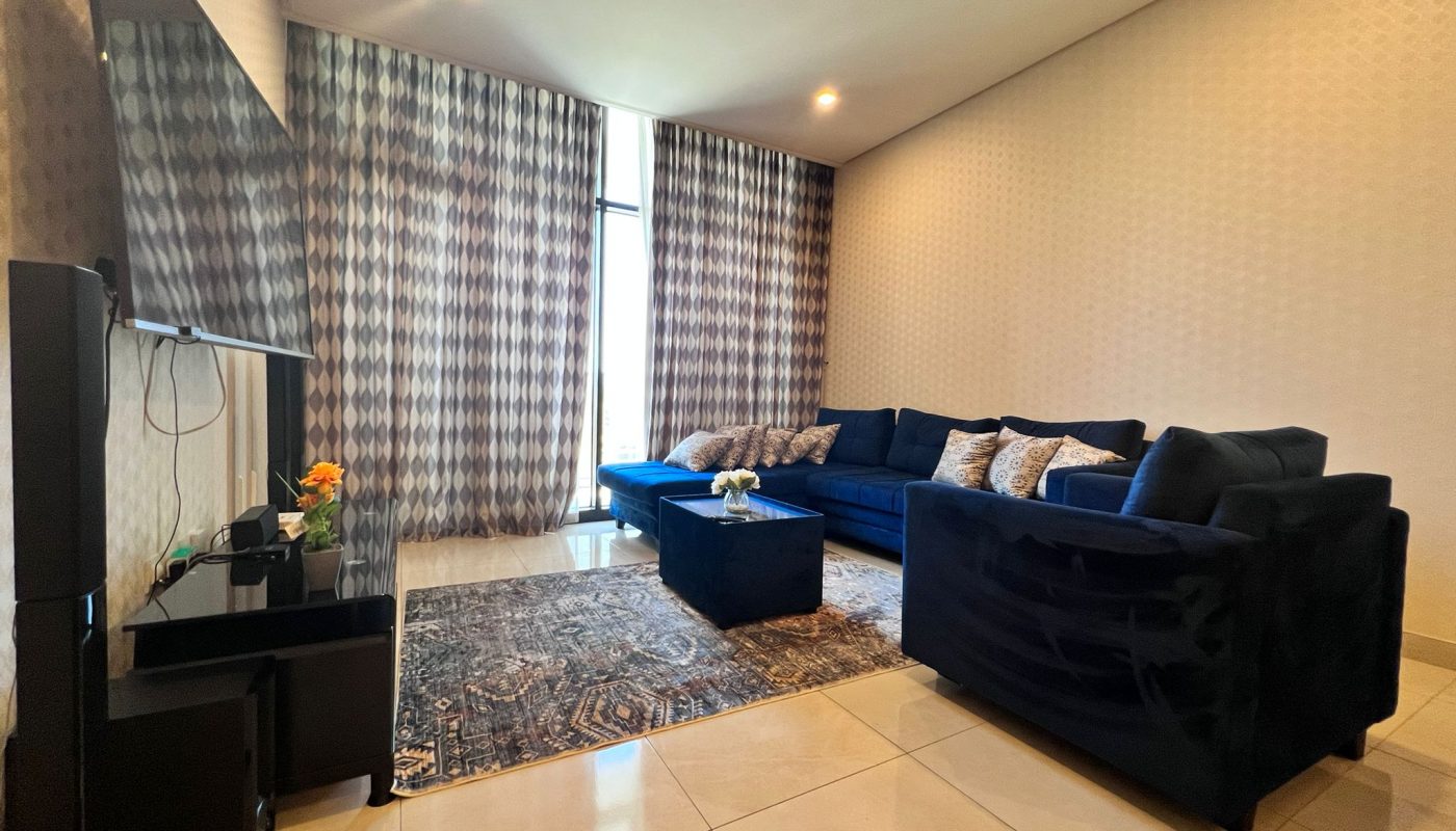 Modern living room with dark blue sectional sofa, patterned rug, TV on a stand, glass coffee table, and large windows with geometric patterned curtains.