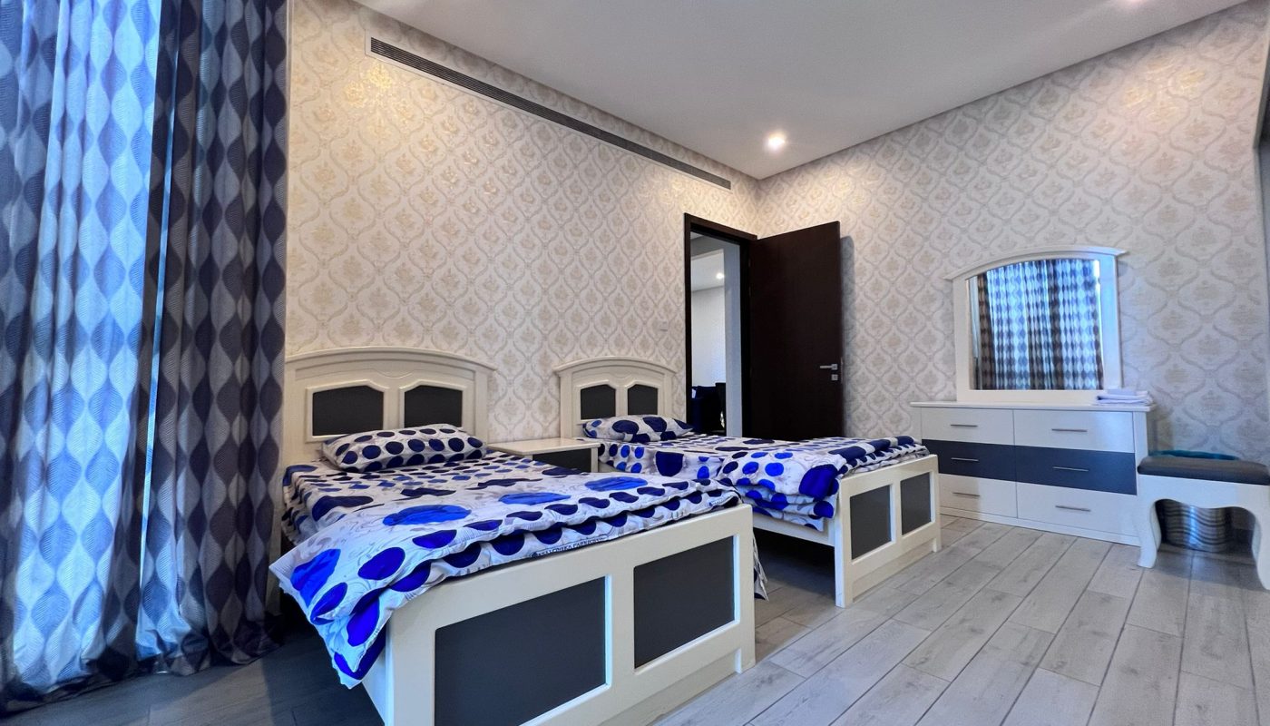 A bedroom with two single beds, a dresser with a mirror, patterned wallpaper, and blue and white bedding and curtains.