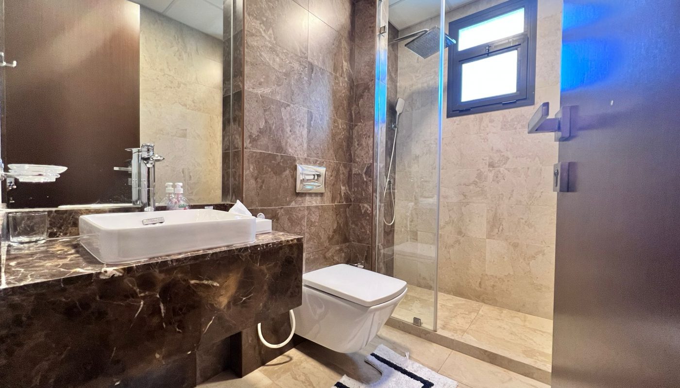 Modern bathroom with marble countertop, vessel sink, wall-mounted toilet, walk-in glass shower, and small window. White bath mat on tiled floor.