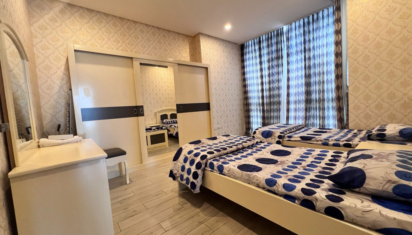 A modern bedroom with two single beds, a dressing table with a mirror, a stool, sliding-door wardrobe, and large window with patterned curtains.