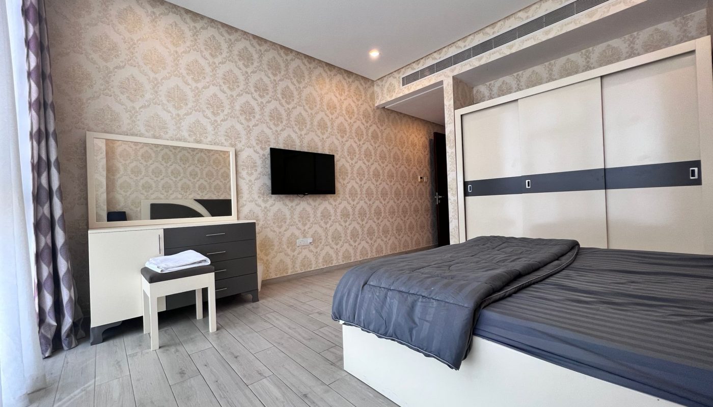 Modern bedroom with patterned wallpaper, a bed with gray bedding, a dresser with a mirror, a small stool, a television mounted on the wall, and a sliding door wardrobe.