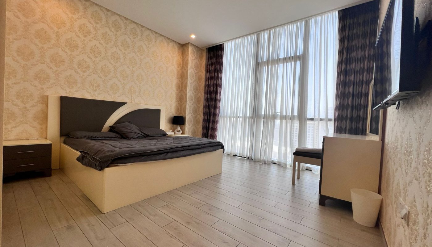 Modern bedroom with a double bed, two nightstands, a desk and chair, large windows with sheer and dark curtains, and light wood flooring.