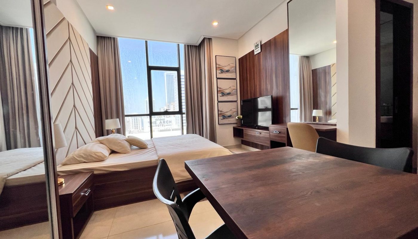 Modern studio apartment with a bed, wooden table and chairs, TV, large window with city view, and mirrored closet doors.