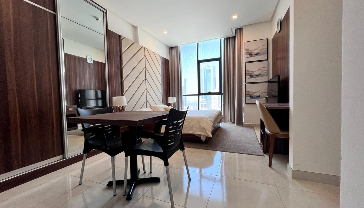 Modern studio apartment with a bed, dining table, TV, desk, large window, and wood-paneled accents. Light-colored floor tiles and wall art add to the contemporary decor.