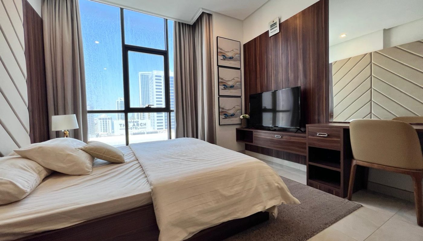 Modern bedroom with large window, double bed, desk, wall-mounted TV, and neutral decor. City buildings visible outside.