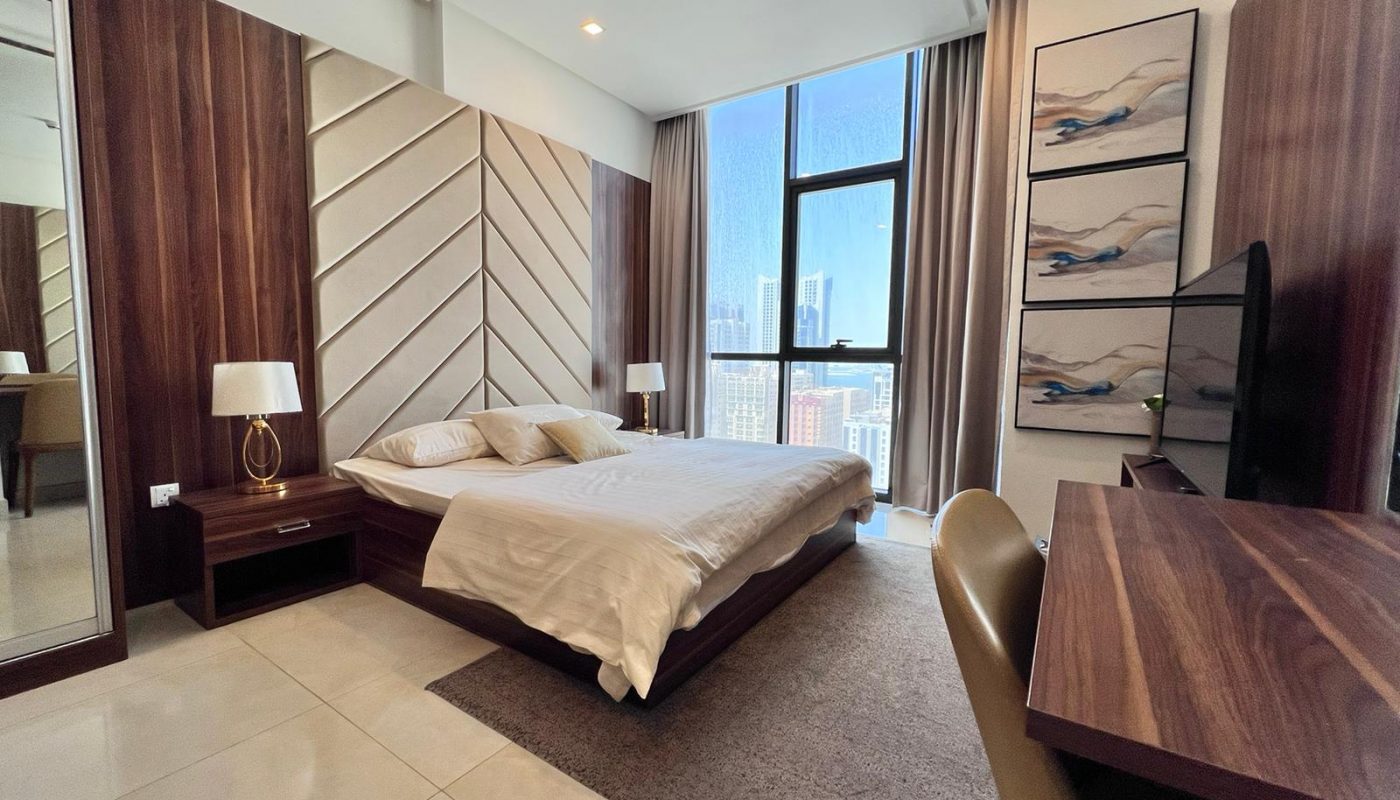 Modern bedroom with large windows, a double bed, wooden furniture, a desk, wall art, and cityscape views outside. Natural light fills the room.