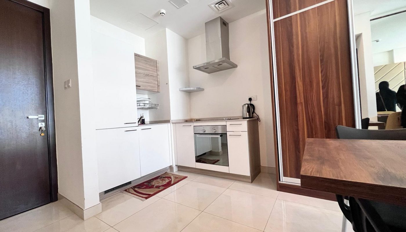 Modern kitchen with white cabinets, built-in oven, stovetop, stainless steel hood, kettle, and a wooden wardrobe. Tile flooring and a dining table with chairs are visible.