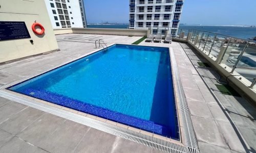 A spacious swimming pool on top of a fully furnished building with a view of the ocean.