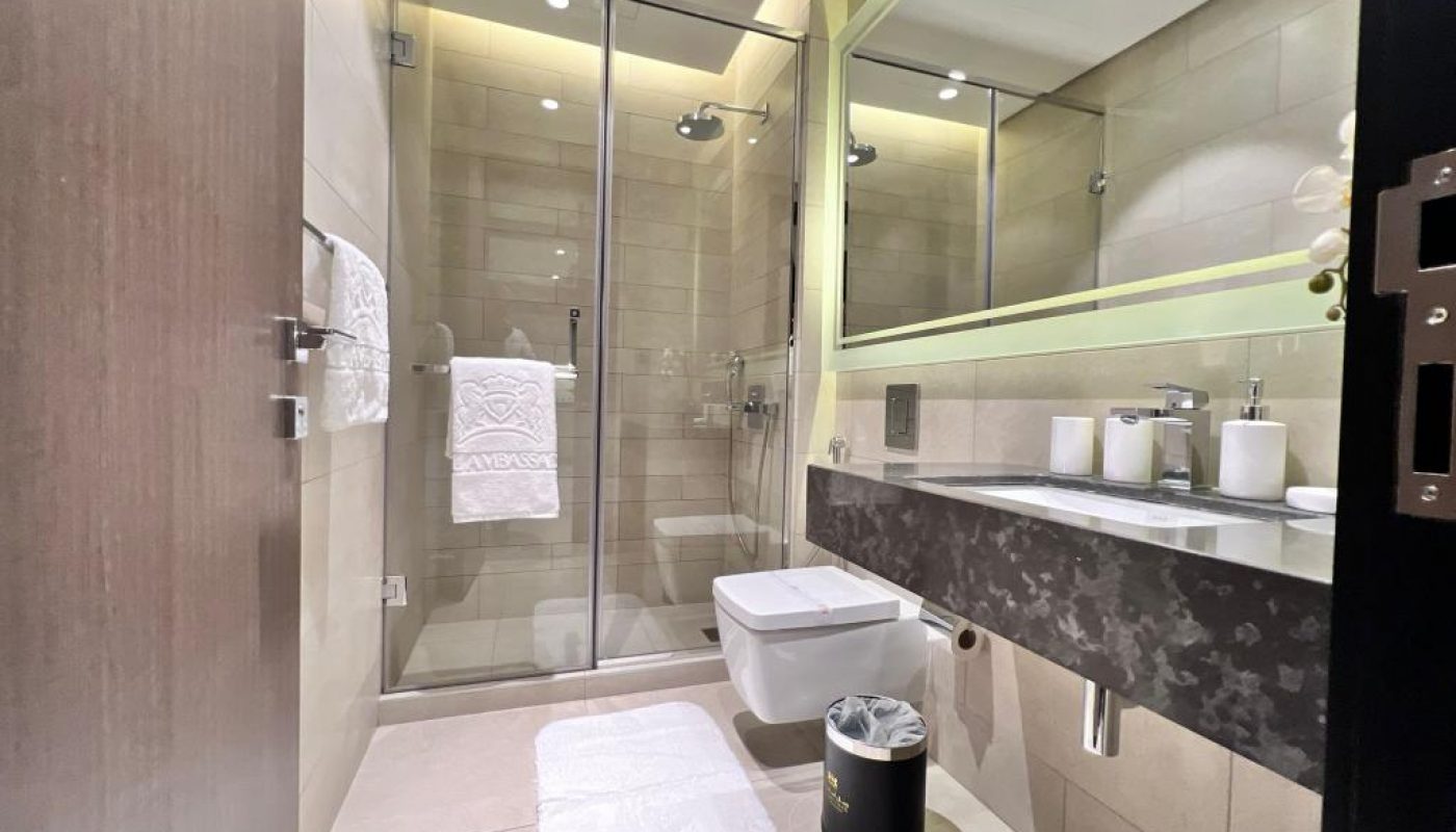 Modern bathroom with a large glass shower, wall-mounted toilet, wide sink with double faucets, countertop amenities, towel rack, white bath mat, and small trash bin.