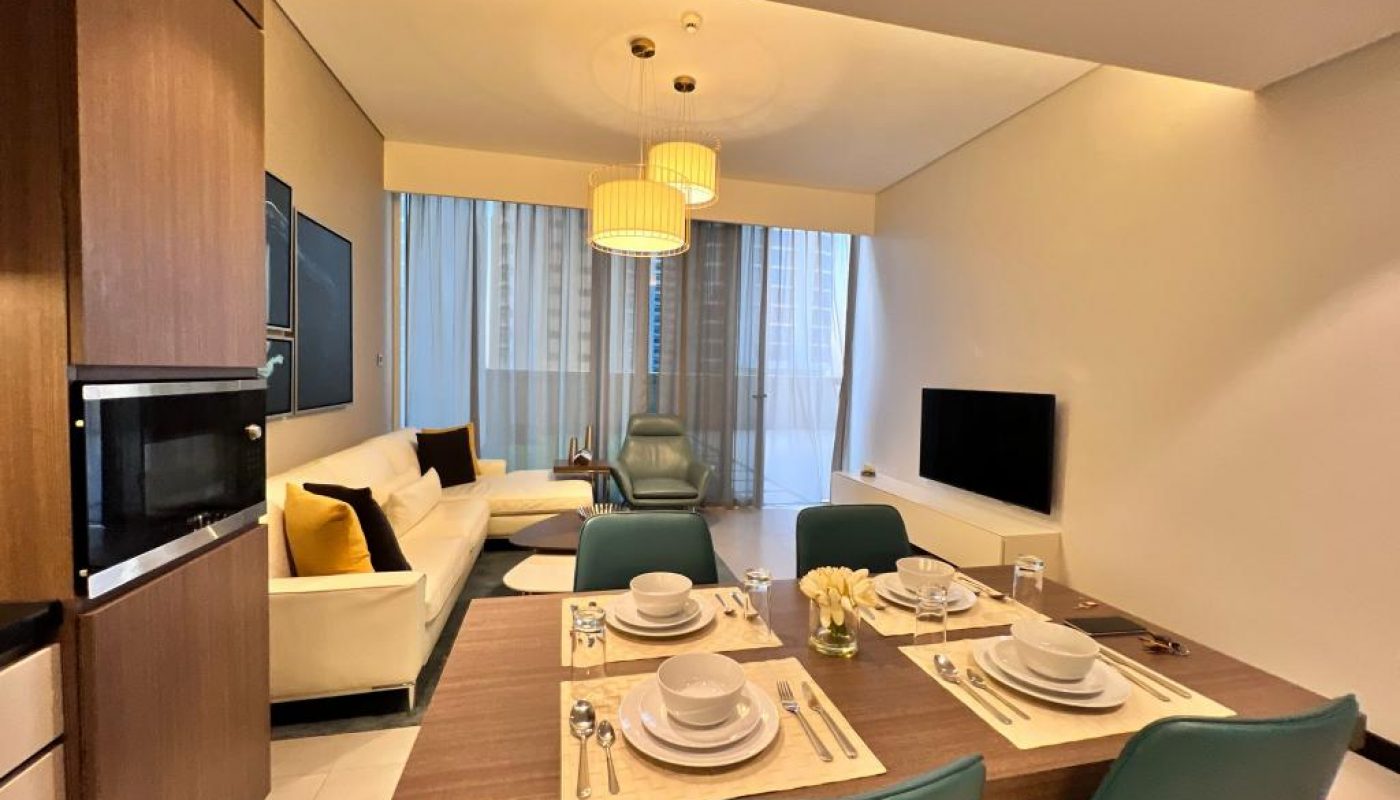 Modern living and dining area with a set dining table, teal chairs, a white sofa, a lounge chair, wall-mounted TV, and large windows with city views.