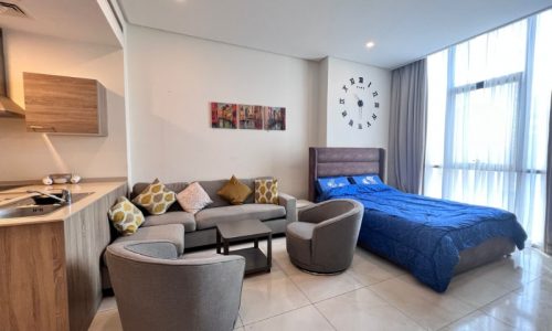 Modern flat for sale in Juffair with a combined living and sleeping area, featuring a large bed, a sofa with decorative pillows, an armchair, wall art, and an oversized wall clock.