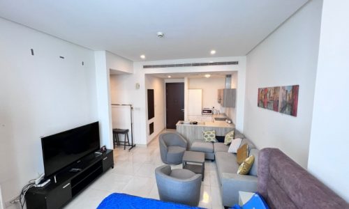 Modern studio flat for sale in Juffair with a combined living room and bedroom space, featuring a couch, bed, television, and kitchenette in the background.