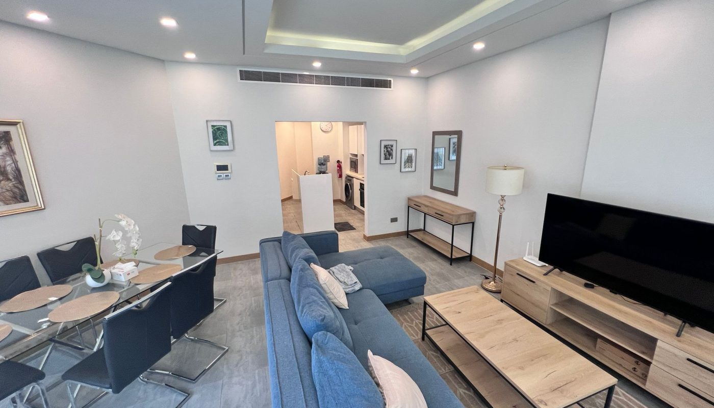 Modern living room with a blue sectional sofa, wooden furniture, dining table with six chairs, large TV, and neutral decor. A kitchen area is visible through an open doorway in the background.