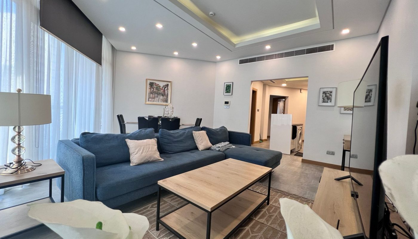Modern living room with blue sectional sofa, wooden coffee table, floor lamp, large TV, dining area, and artwork on white walls. Natural light streams in through sheer curtains.