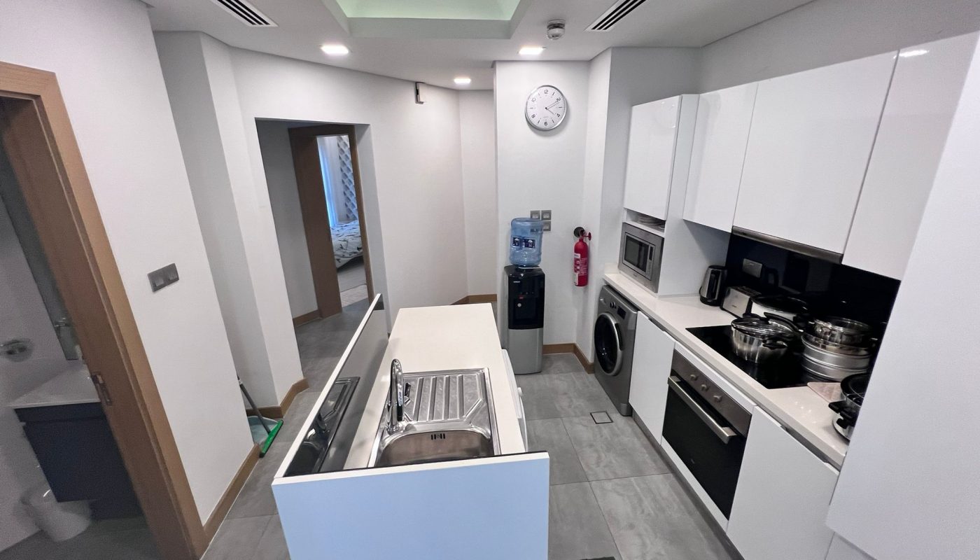 Modern white kitchen with built-in appliances, a sink island, a wall clock, and a water dispenser. Doorways lead to a bathroom and another room. Pots and pans sit on the stovetop.
